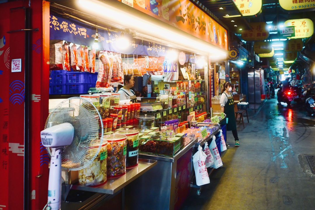 宜蘭傳統市場內的熱鬧街景，畫面左側為知名的三源臘味行乾貨攤位，販售鴨賞與臘肉等在地名產，展現道地的宜蘭迺菜市場氛圍