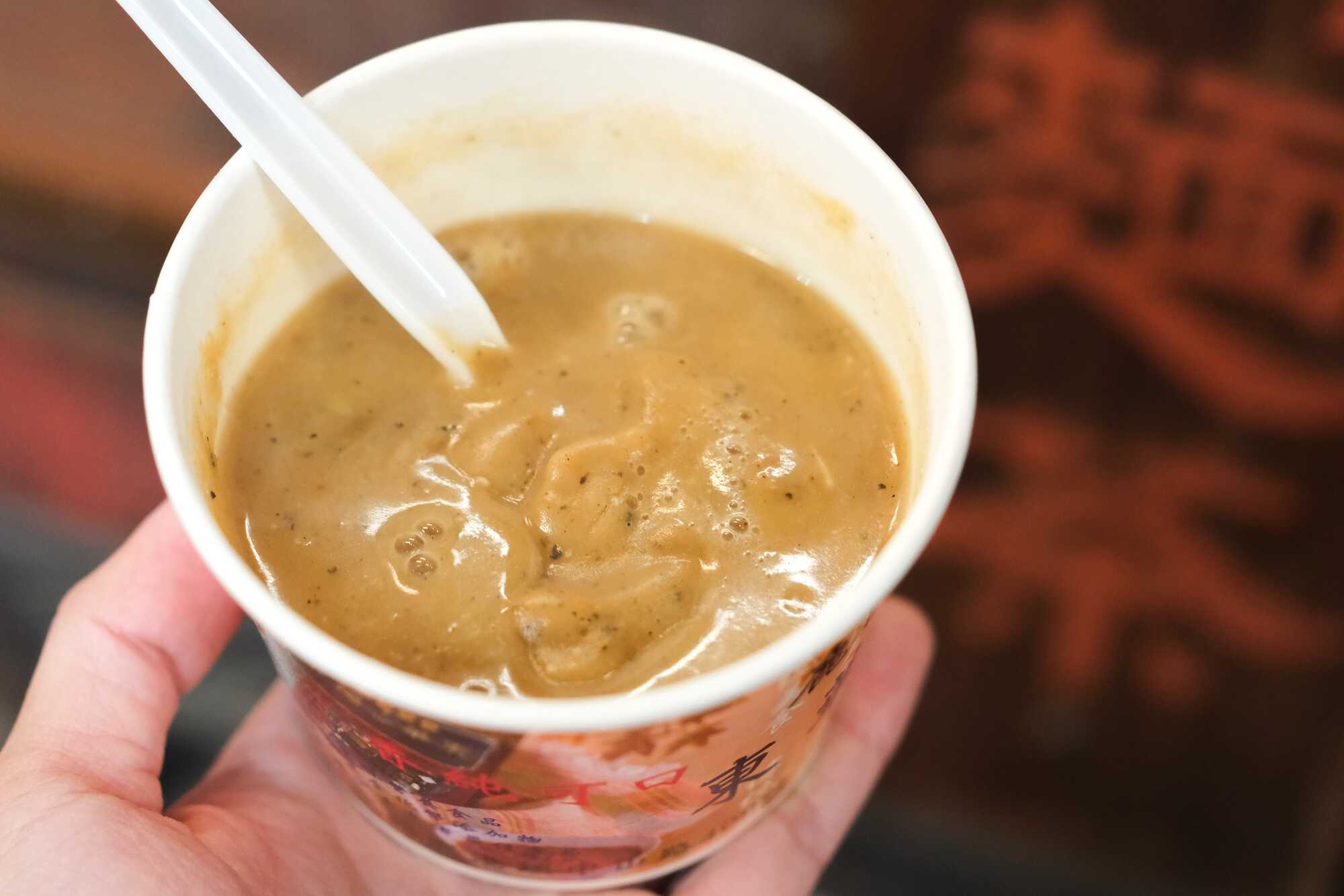 手持一杯濃郁的鹿港傳統美食熱麵茶,杯中插著湯匙,背景隱約可見麵茶攤位的字樣。